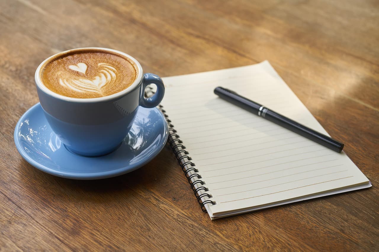 Heart latte art in a blue cup beside a notebook and pen.