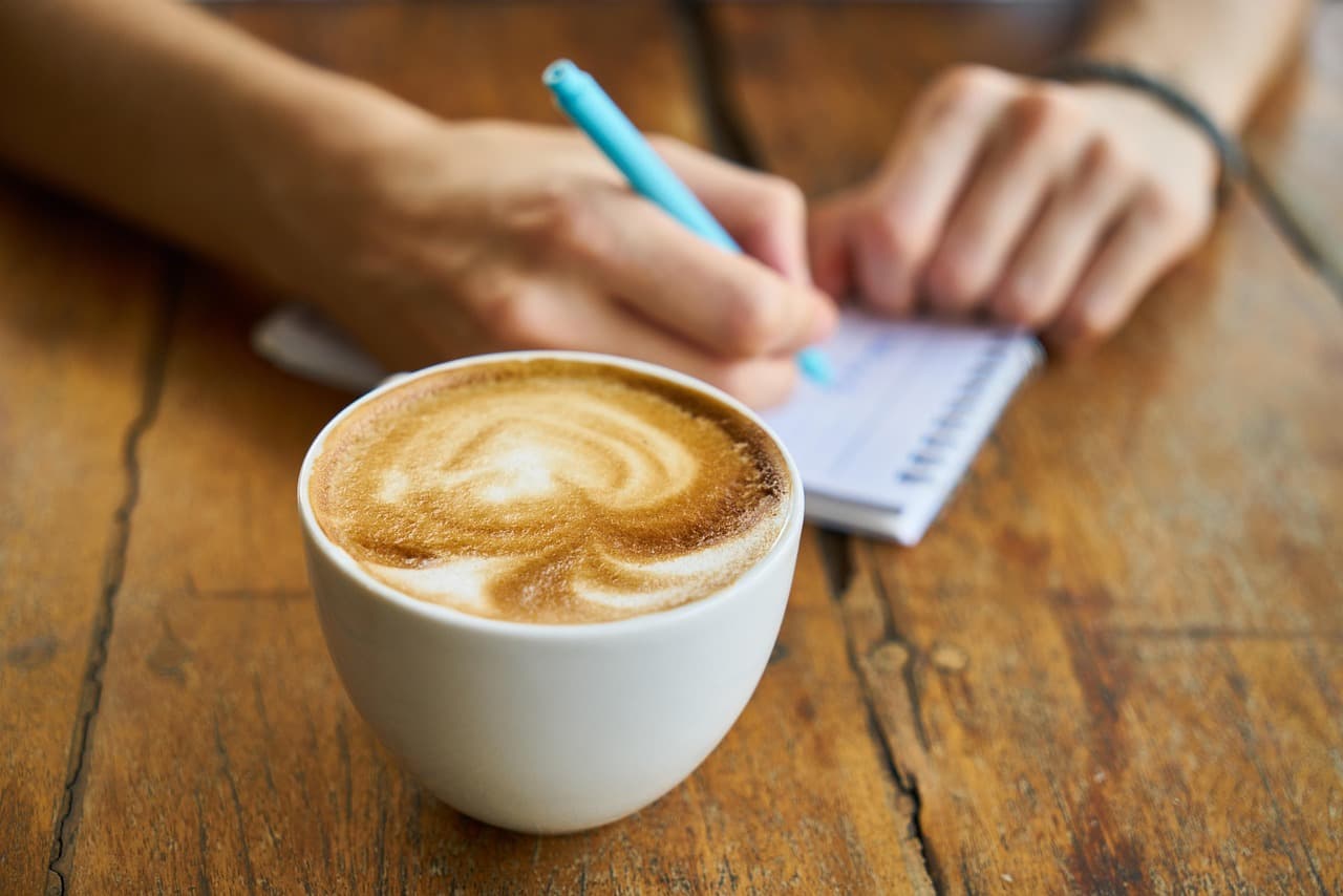 A latte on a worn wood table while someone writes in a notebook.